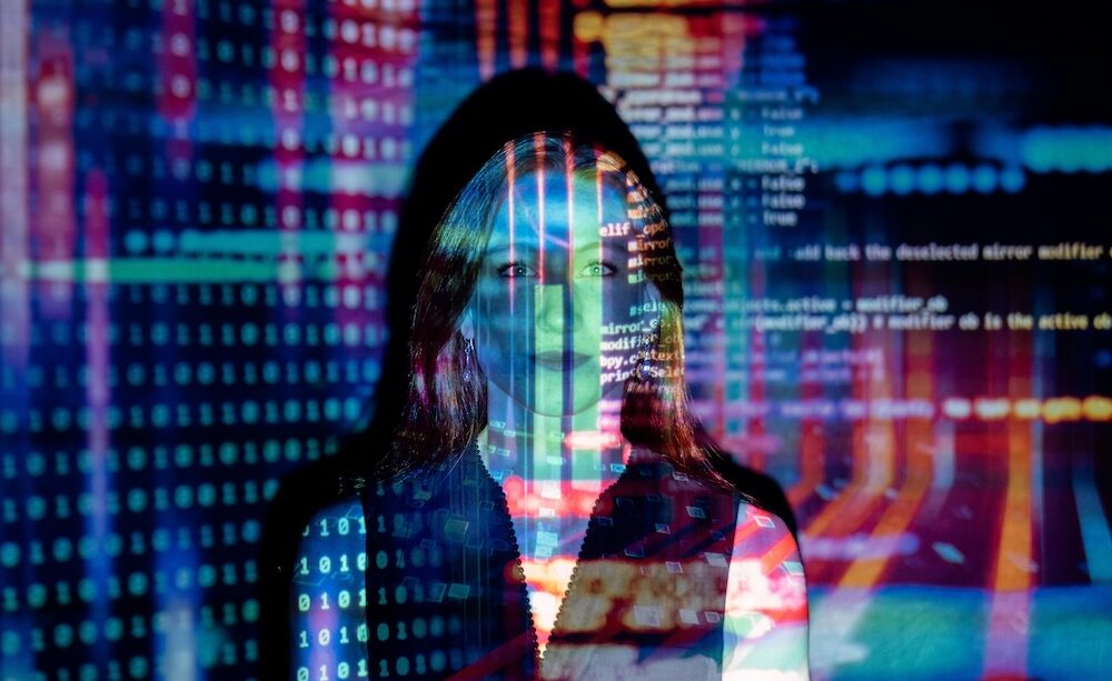 A woman with long hair stands in front of a digital projection featuring binary code, data streams, and colorful programming code. This visually represents AI-powered marketing, automation, and data-driven decision-making—key themes in the blog discussing how Robotic Marketer streamlines strategy, execution, and reporting using artificial intelligence.